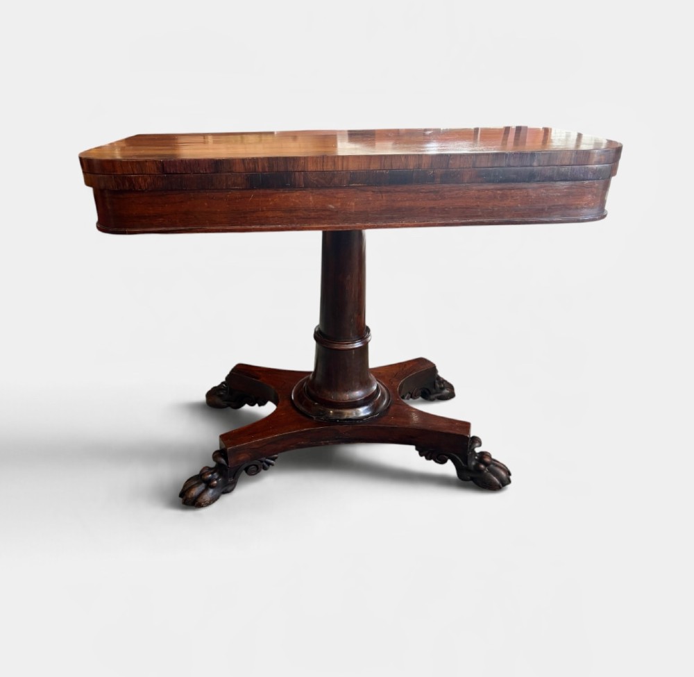 A William IV rosewood fold-over card table featuring a cylindrical central pedestal resting on a concave four-point base with carved lion claw feet.