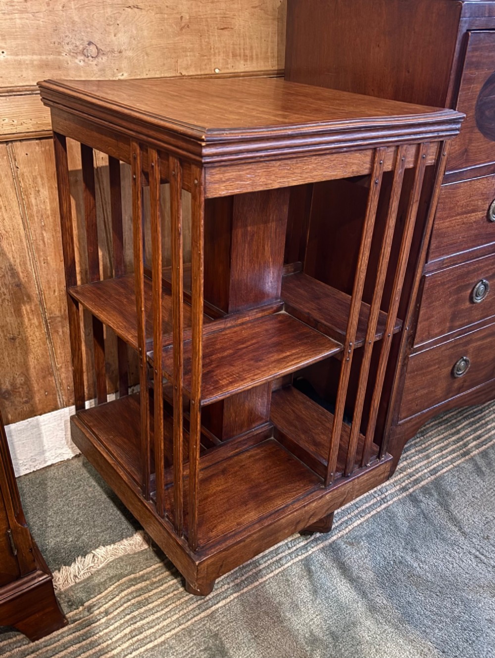 An Edwardian oak revolving bookcase featuring two tiers with vertical slatted sides mounted on a rotating central base. View IV