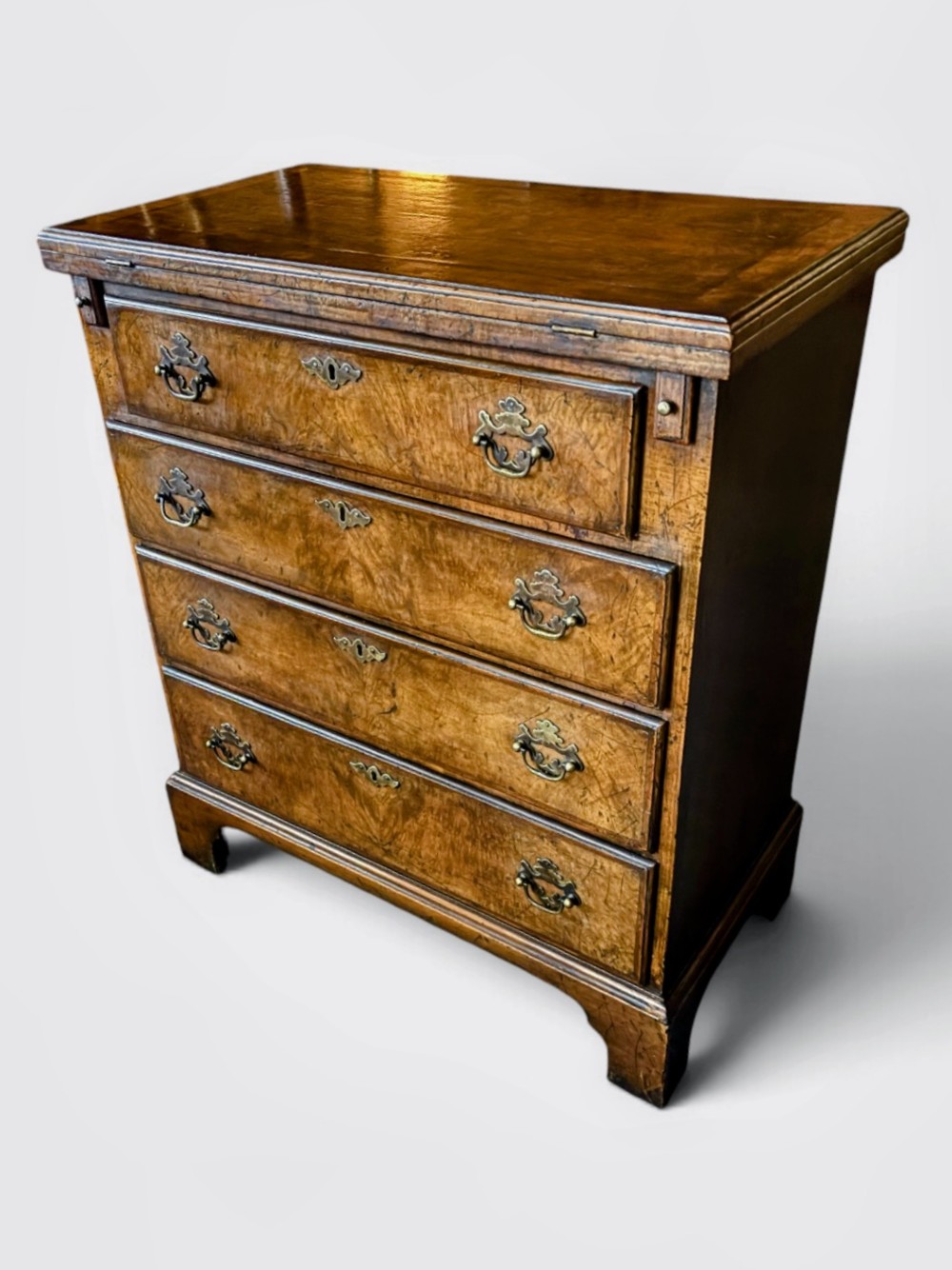 An antique burr walnut bachelors chest featuring a hinged fold-over top, four graduated drawers with brass handles, and bracket feet. View IV
