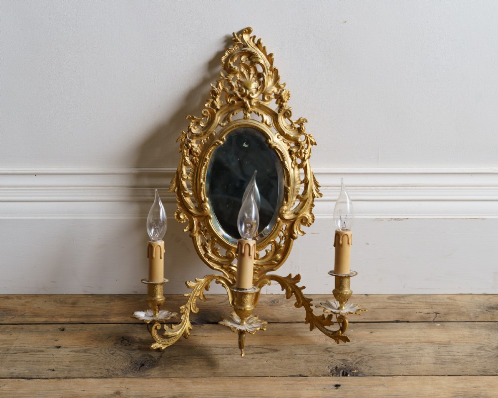 An ornate Rococo style gilt metal girandole featuring a central oval mirror surrounded by scrollwork and three candle-effect light branches.
