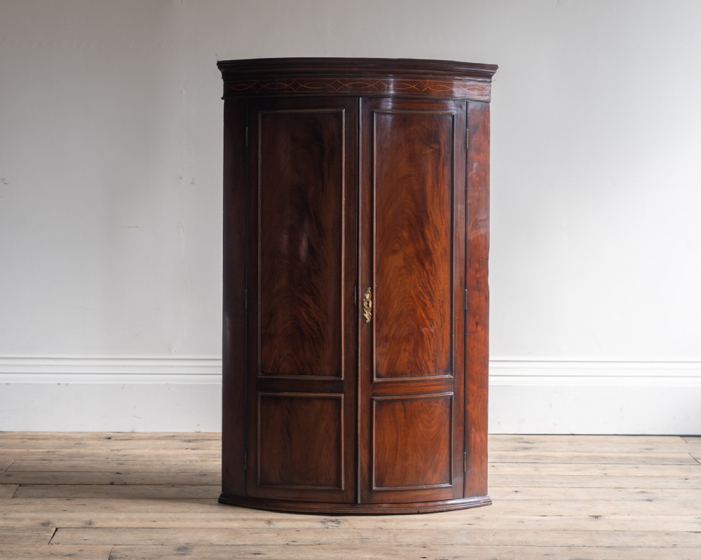 A Georgian mahogany bowfront corner cupboard featuring twin doors with flame mahogany veneer panels, an inlaid upper frieze, and a brass escutcheon.