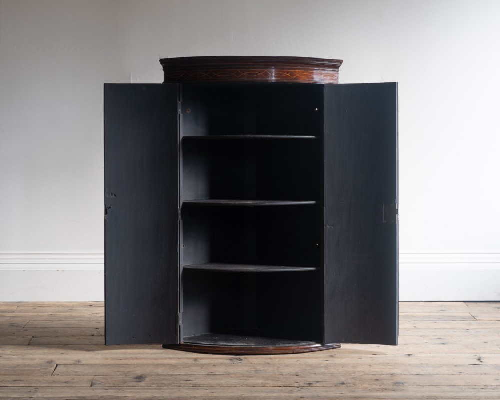 A Georgian mahogany bowfront corner cupboard featuring twin doors with flame mahogany veneer panels, an inlaid upper frieze, and a brass escutcheon. View II