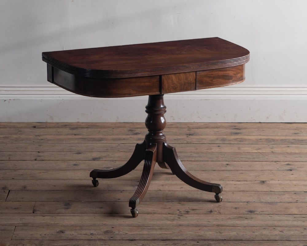 A dark mahogany Regency fold-over tea table featuring a curved top, central turned pedestal, and four reeded sabre legs with brass castors.