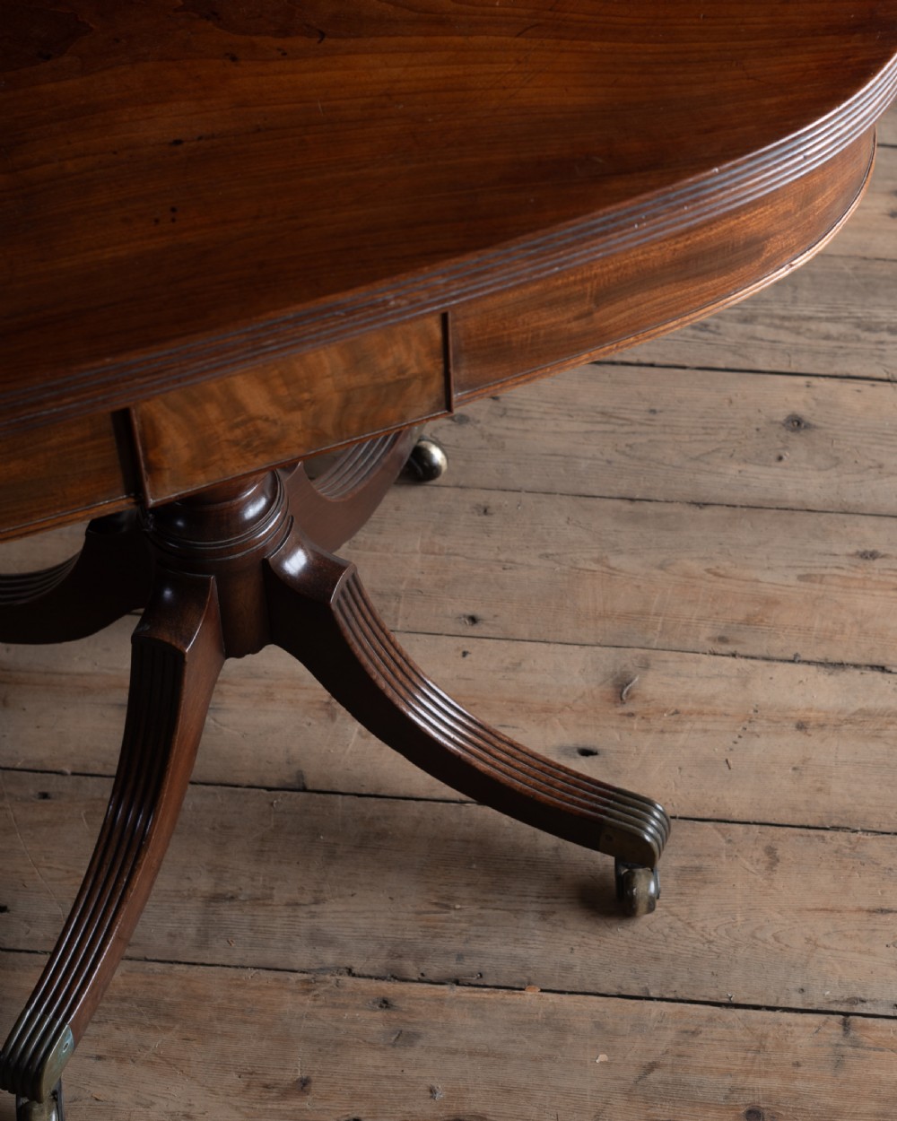 A dark mahogany Regency fold-over tea table featuring a curved top, central turned pedestal, and four reeded sabre legs with brass castors. View II