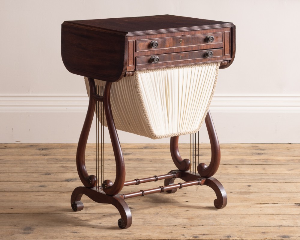 A Regency mahogany sewing table featuring drop leaves, two shallow drawers, a pleated fabric work basket, and lyre-shaped twin supports resting on splayed legs with brass castors.