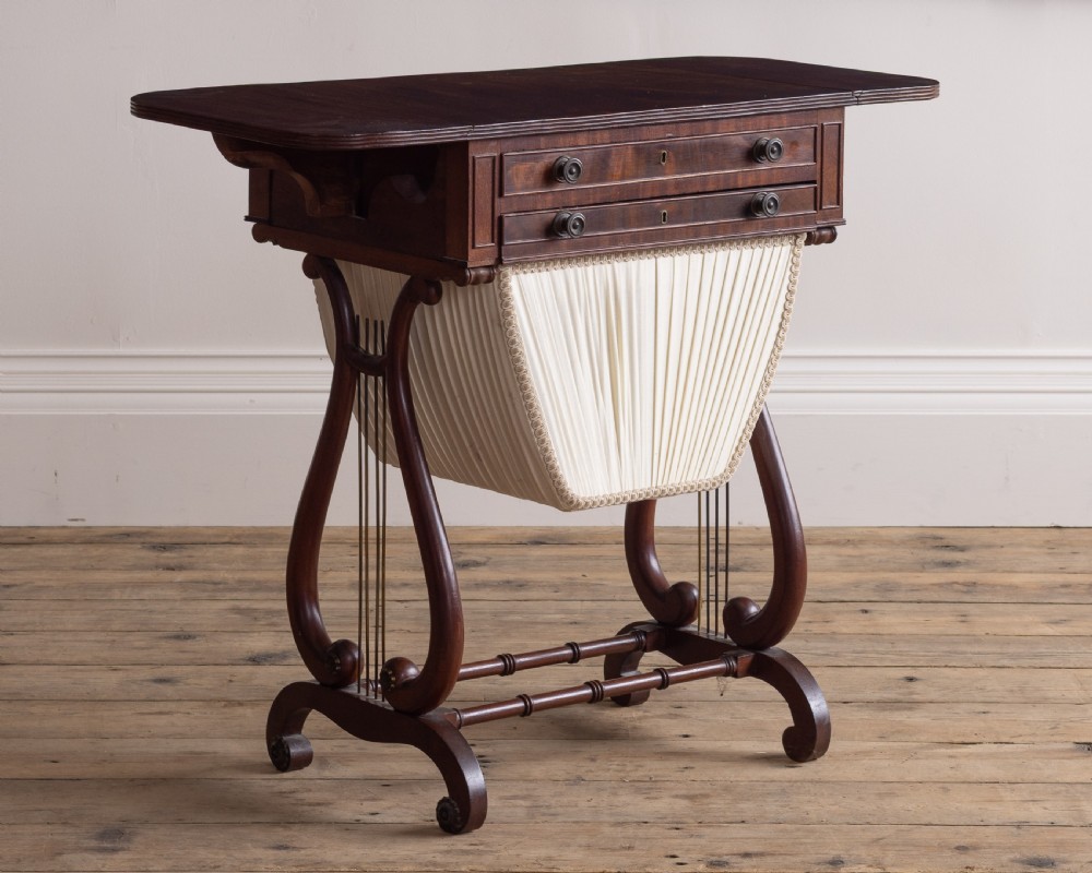 A Regency mahogany sewing table featuring drop leaves, two shallow drawers, a pleated fabric work basket, and lyre-shaped twin supports resting on splayed legs with brass castors. View II
