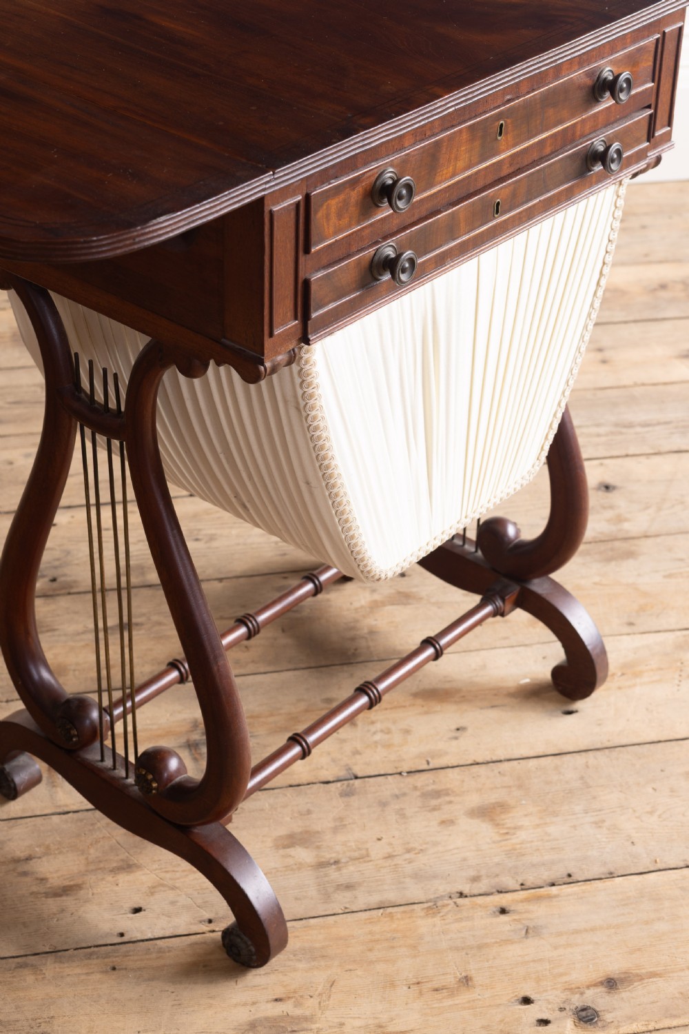 A Regency mahogany sewing table featuring drop leaves, two shallow drawers, a pleated fabric work basket, and lyre-shaped twin supports resting on splayed legs with brass castors. View III