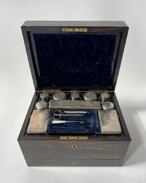 fig 1. A nineteenth-century dark coromandel wood dressing case open to reveal dark blue velvet interior lining, fitted silver-topped glass bottles, vanity implements, and an extended lower drawer.