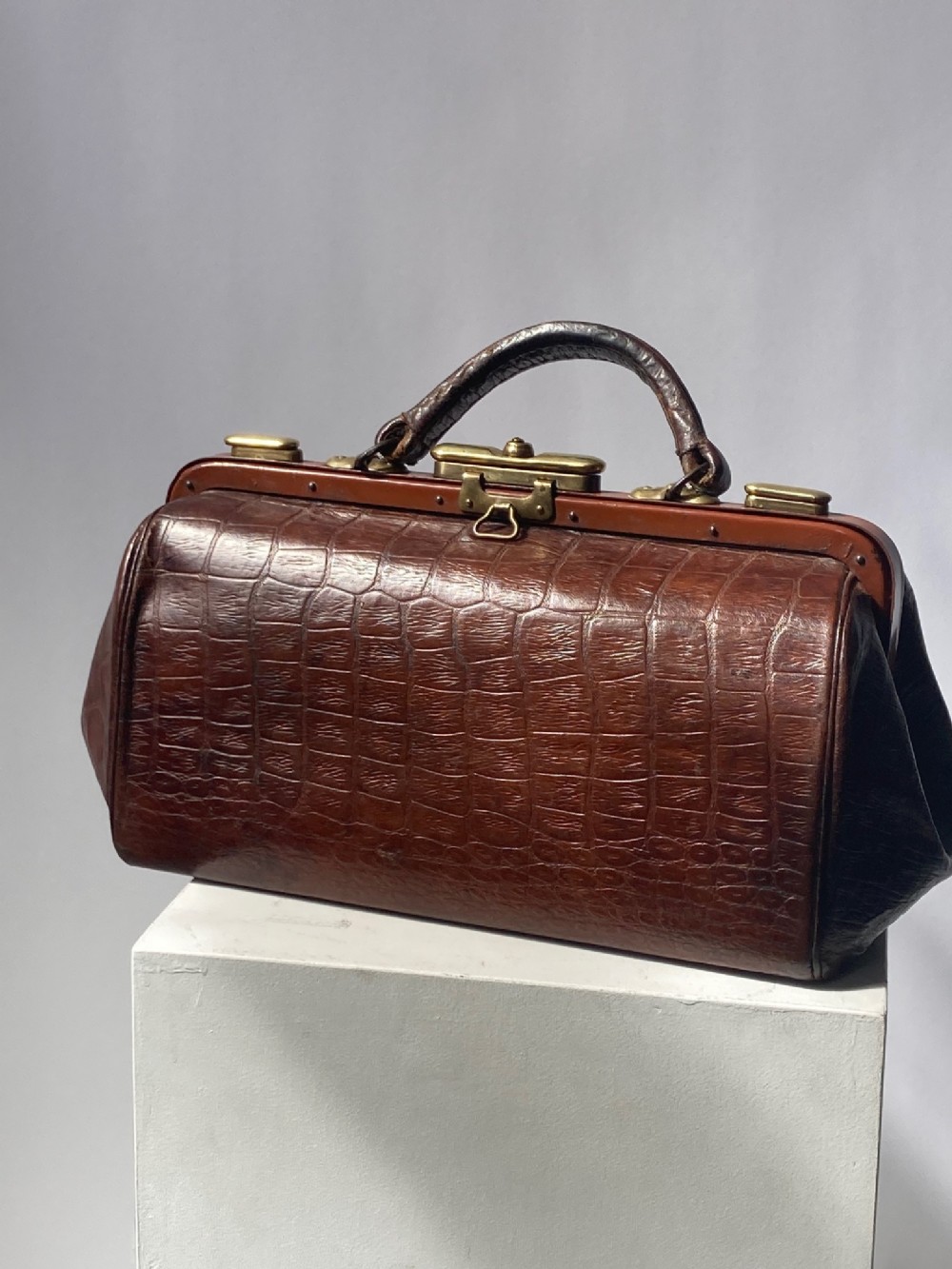 A warm brown leather Gladstone bag with crocodile skin texture, featuring a hinged top frame with brass latches and a top handle.