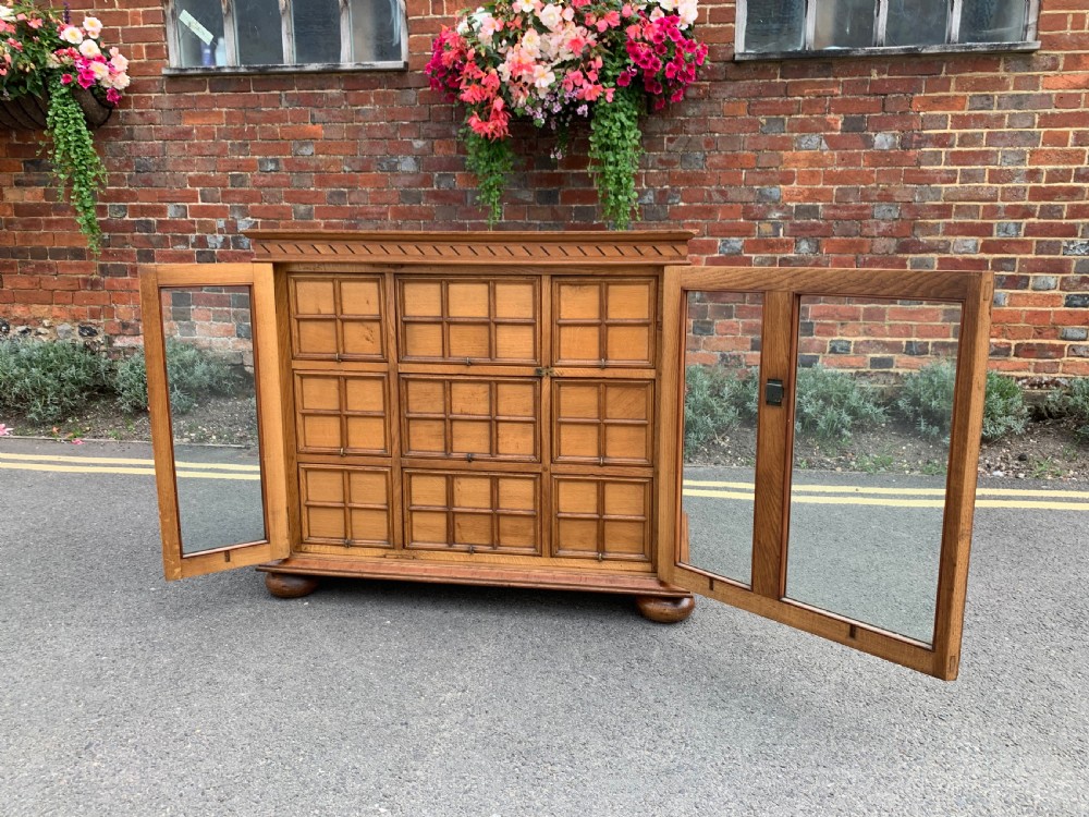 A low three-door oak vestry cupboard featuring clear glass panels, incised frieze carving, and bun feet. View V