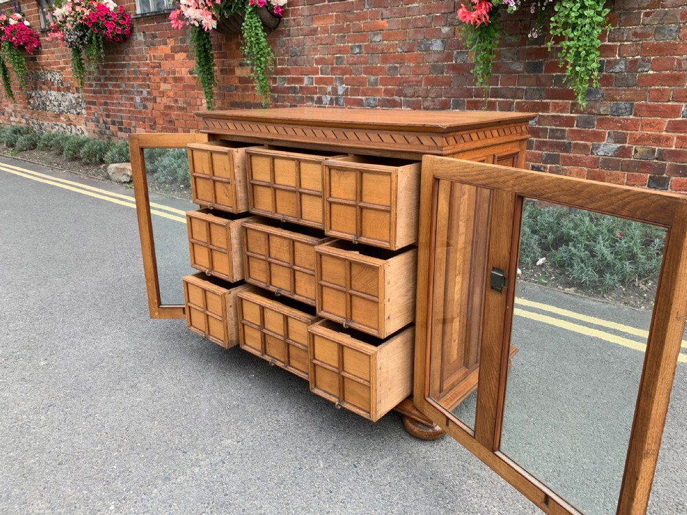 A low three-door oak vestry cupboard featuring clear glass panels, incised frieze carving, and bun feet. View VI