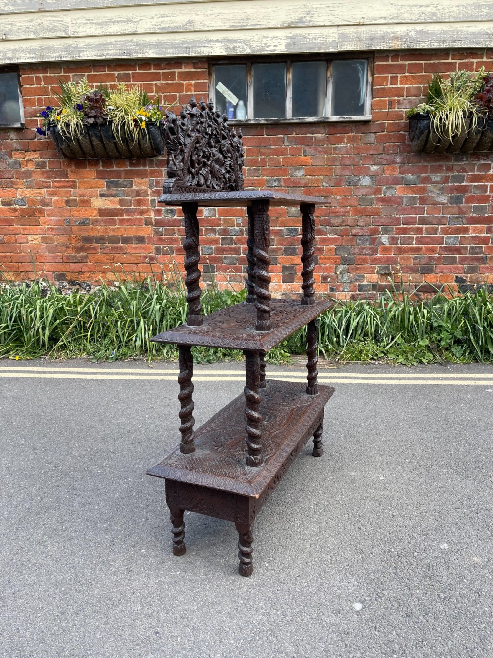 A three-tier dark teak carved whatnot featuring spiralled support posts and a deeply carved mythological cresting gallery at the top. View VI