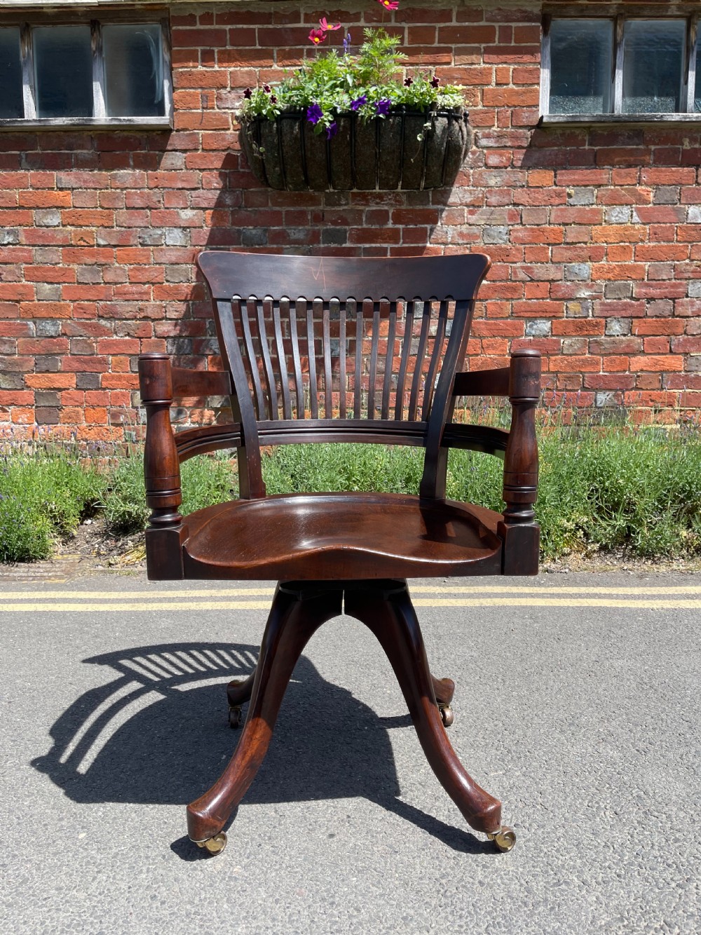 A dark brown wood revolving desk chair featuring a curved slatted backrest, turned arm posts, a contoured seat, and four splayed legs with brass castors.