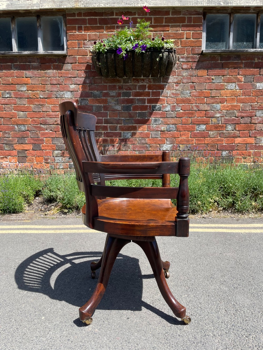 A dark brown wood revolving desk chair featuring a curved slatted backrest, turned arm posts, a contoured seat, and four splayed legs with brass castors. View VI