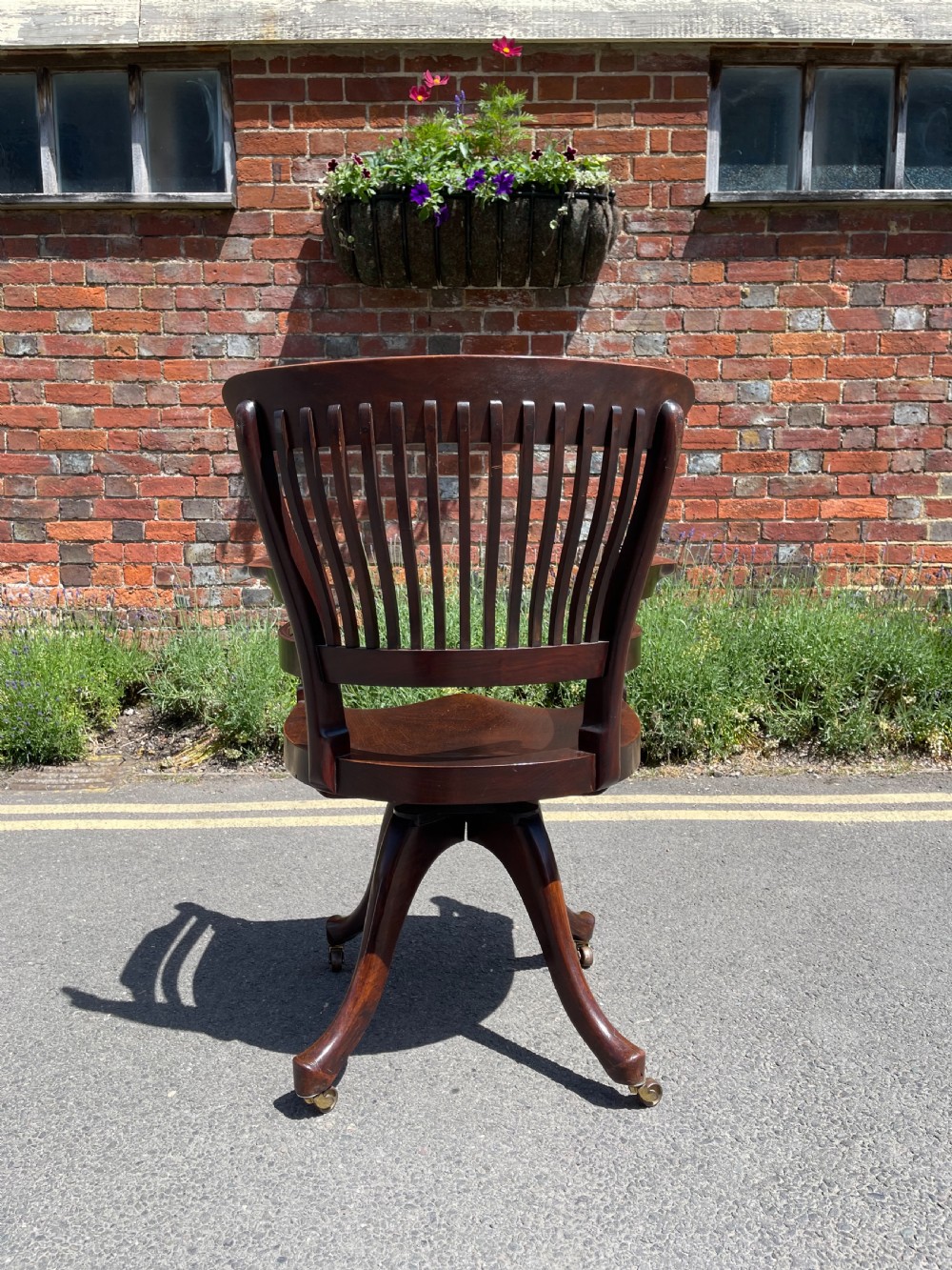A dark brown wood revolving desk chair featuring a curved slatted backrest, turned arm posts, a contoured seat, and four splayed legs with brass castors. View VIII
