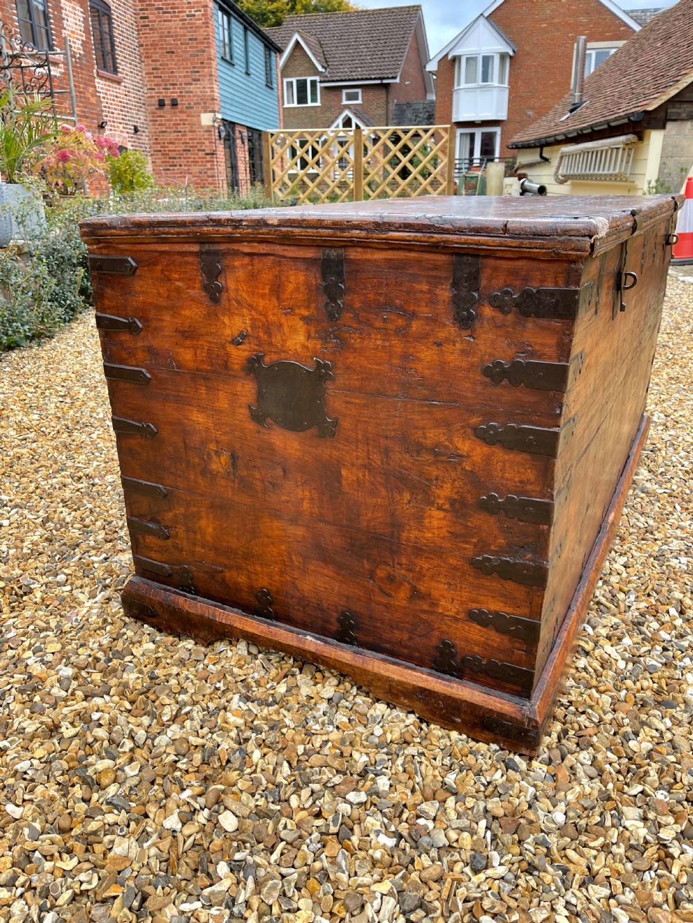 A large warm brown teak storage trunk featuring decorative brass strap hinges and metal corner mounts set on a gravel driveway. View II