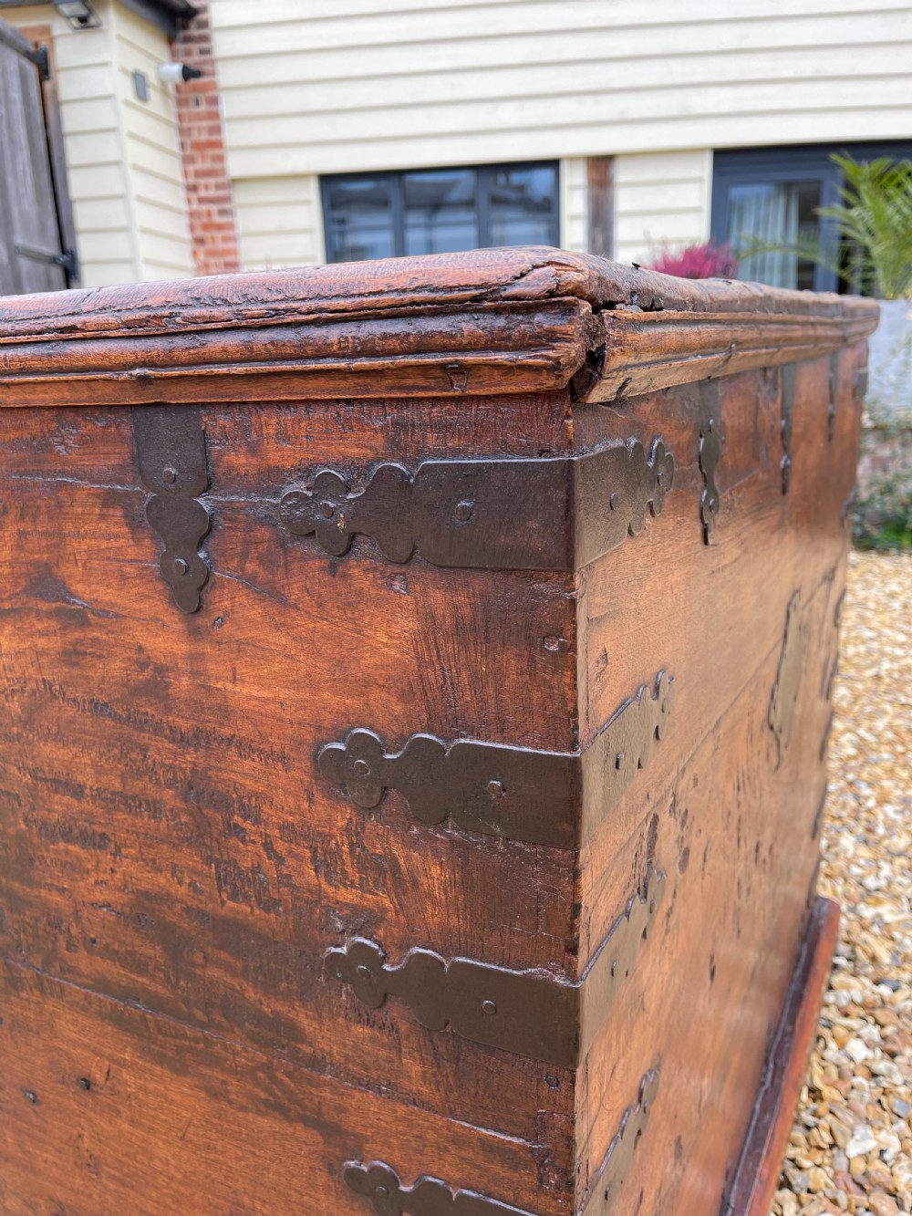 A large warm brown teak storage trunk featuring decorative brass strap hinges and metal corner mounts set on a gravel driveway. View III