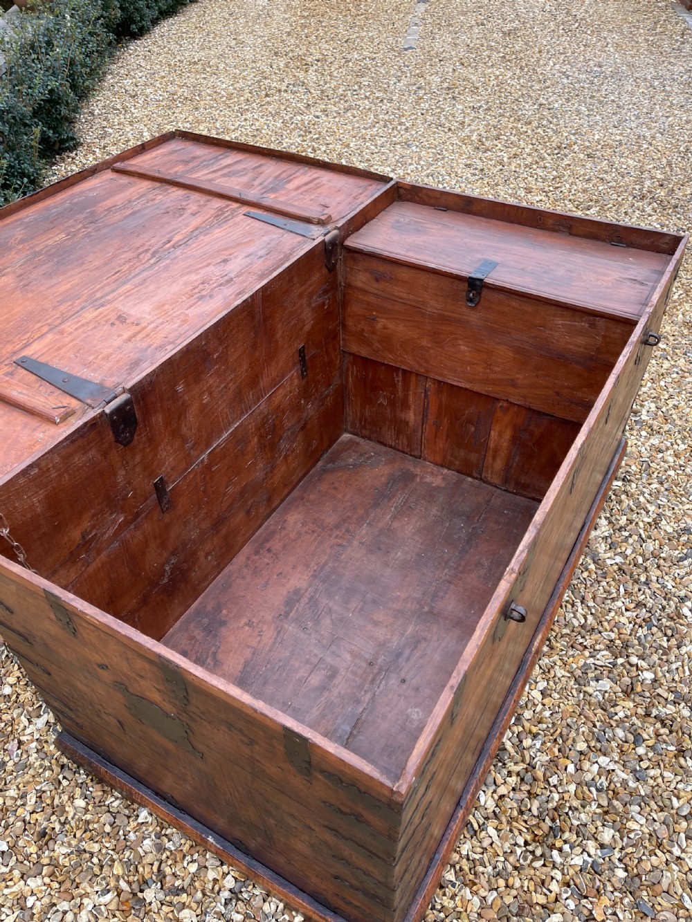 A large warm brown teak storage trunk featuring decorative brass strap hinges and metal corner mounts set on a gravel driveway. View V