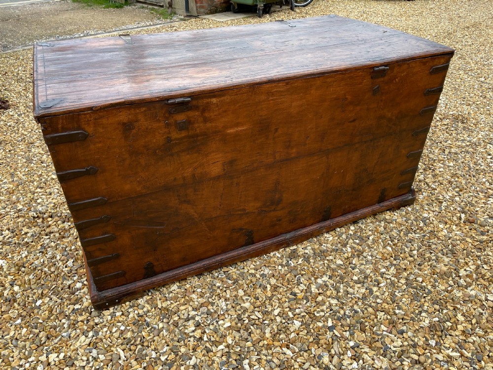 A large warm brown teak storage trunk featuring decorative brass strap hinges and metal corner mounts set on a gravel driveway. View VI