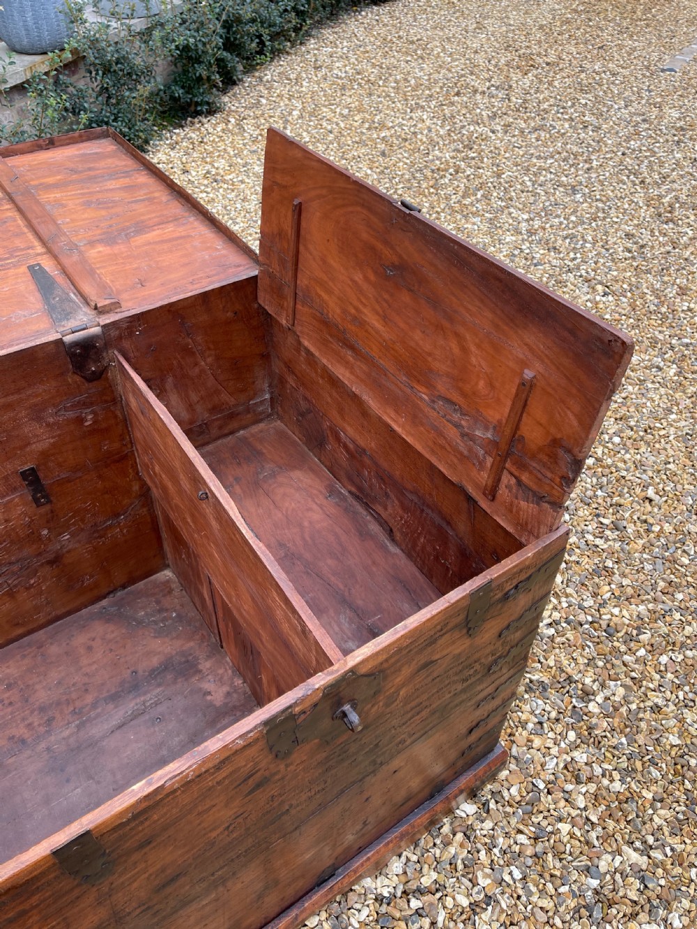 A large warm brown teak storage trunk featuring decorative brass strap hinges and metal corner mounts set on a gravel driveway. View VII