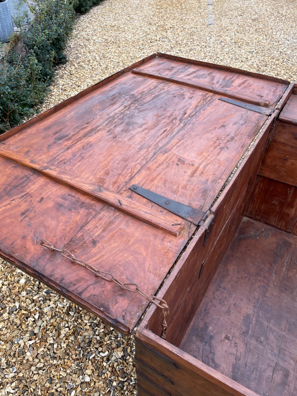 A large warm brown teak storage trunk featuring decorative brass strap hinges and metal corner mounts set on a gravel driveway. View VIII