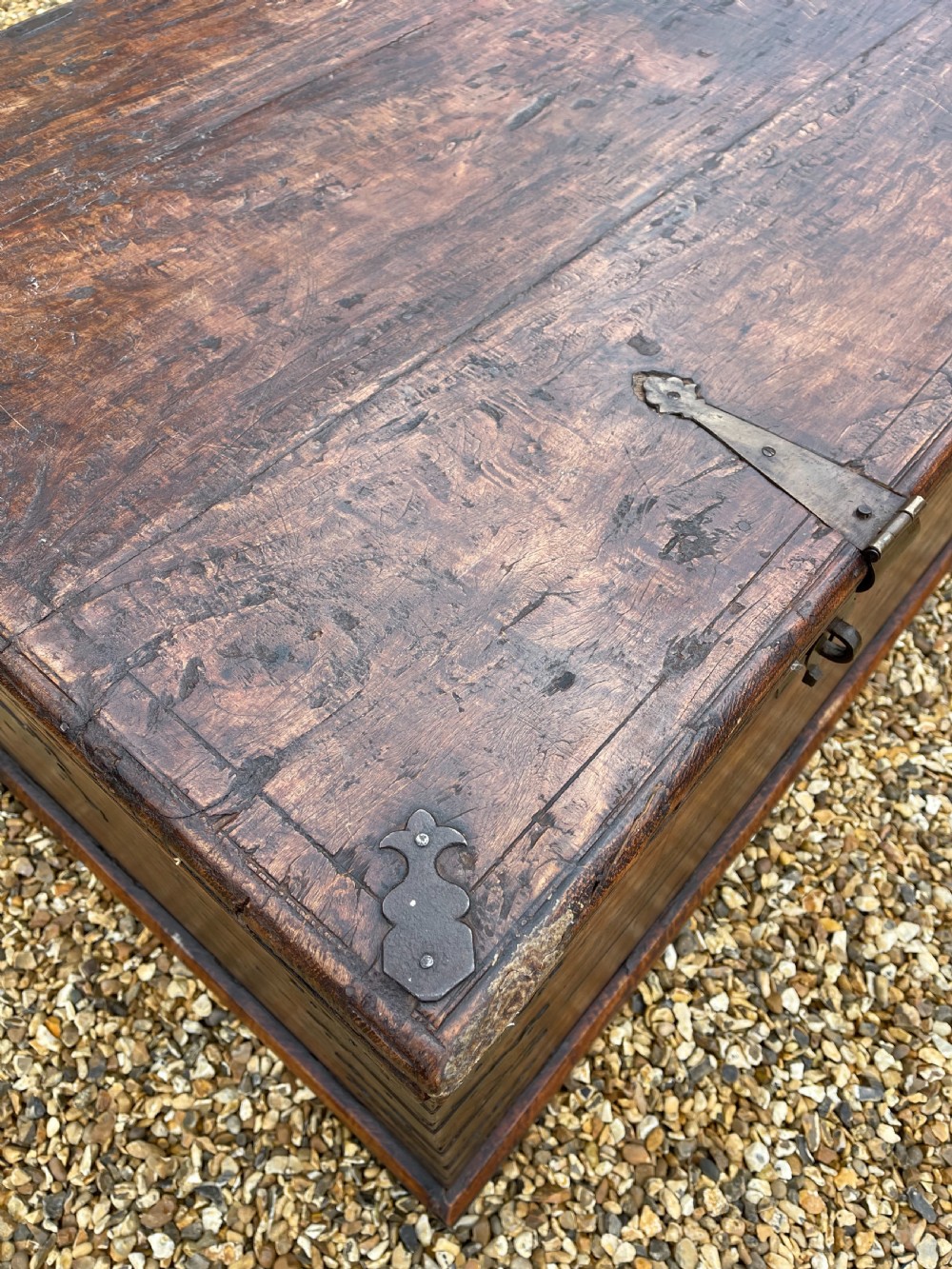 A large warm brown teak storage trunk featuring decorative brass strap hinges and metal corner mounts set on a gravel driveway. View IX