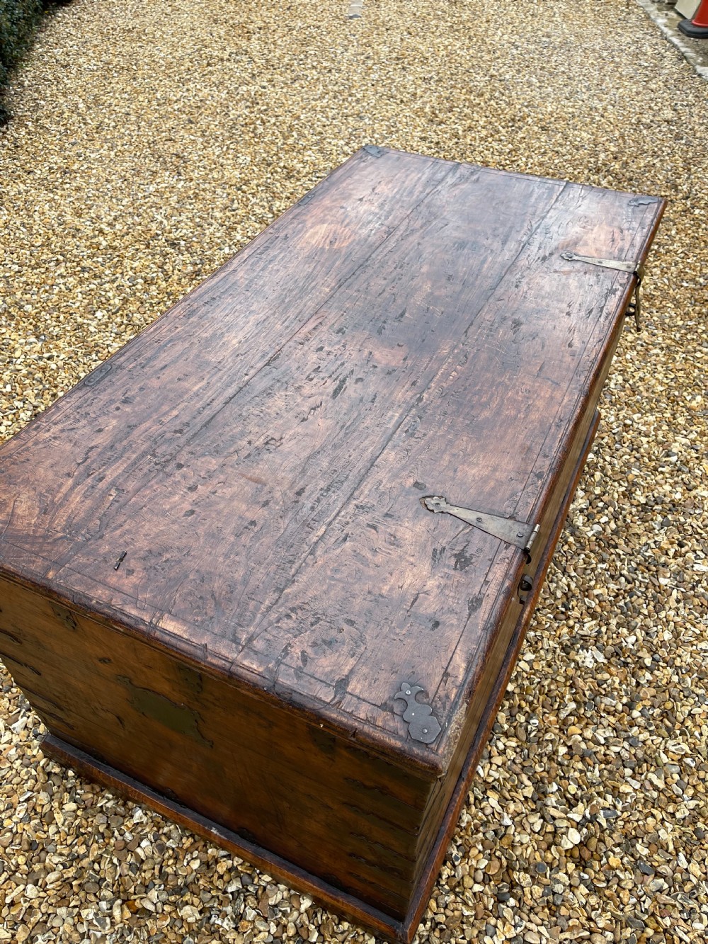 A large warm brown teak storage trunk featuring decorative brass strap hinges and metal corner mounts set on a gravel driveway. View X