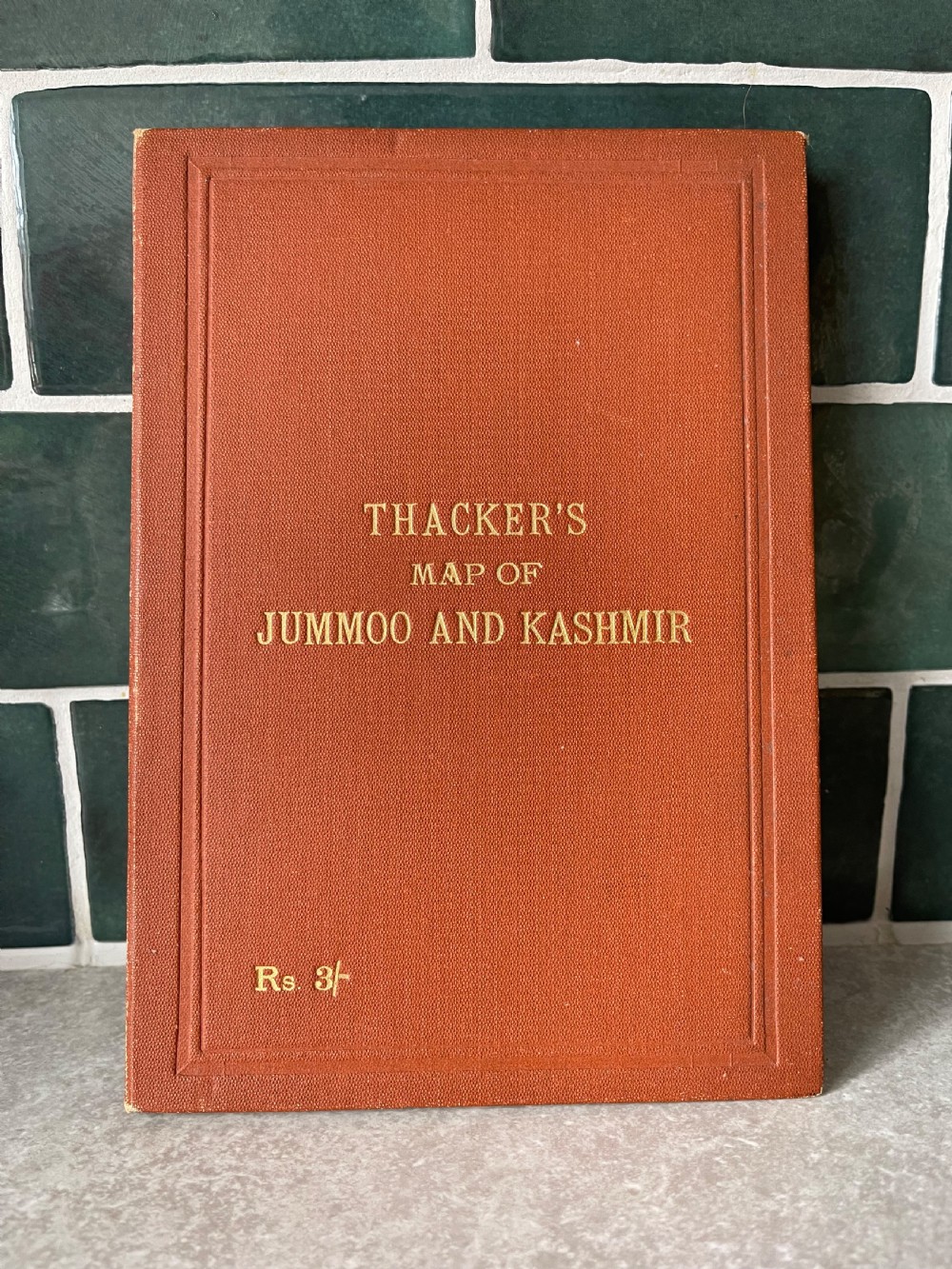 An antique terracotta cloth-bound cover for Thackers Map of Jummoo and Kashmir featuring gilt-embossed lettering and a debossed border.