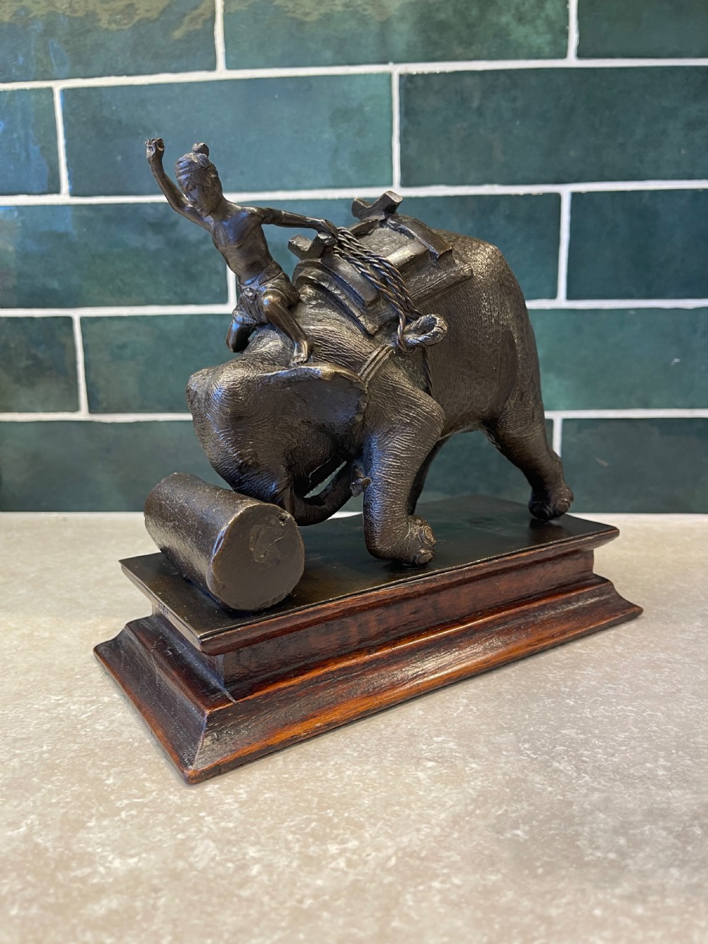 A patinated dark bronze sculpture of a working elephant pushing a log with a mahout riding its neck, mounted on a molded wooden base. View V