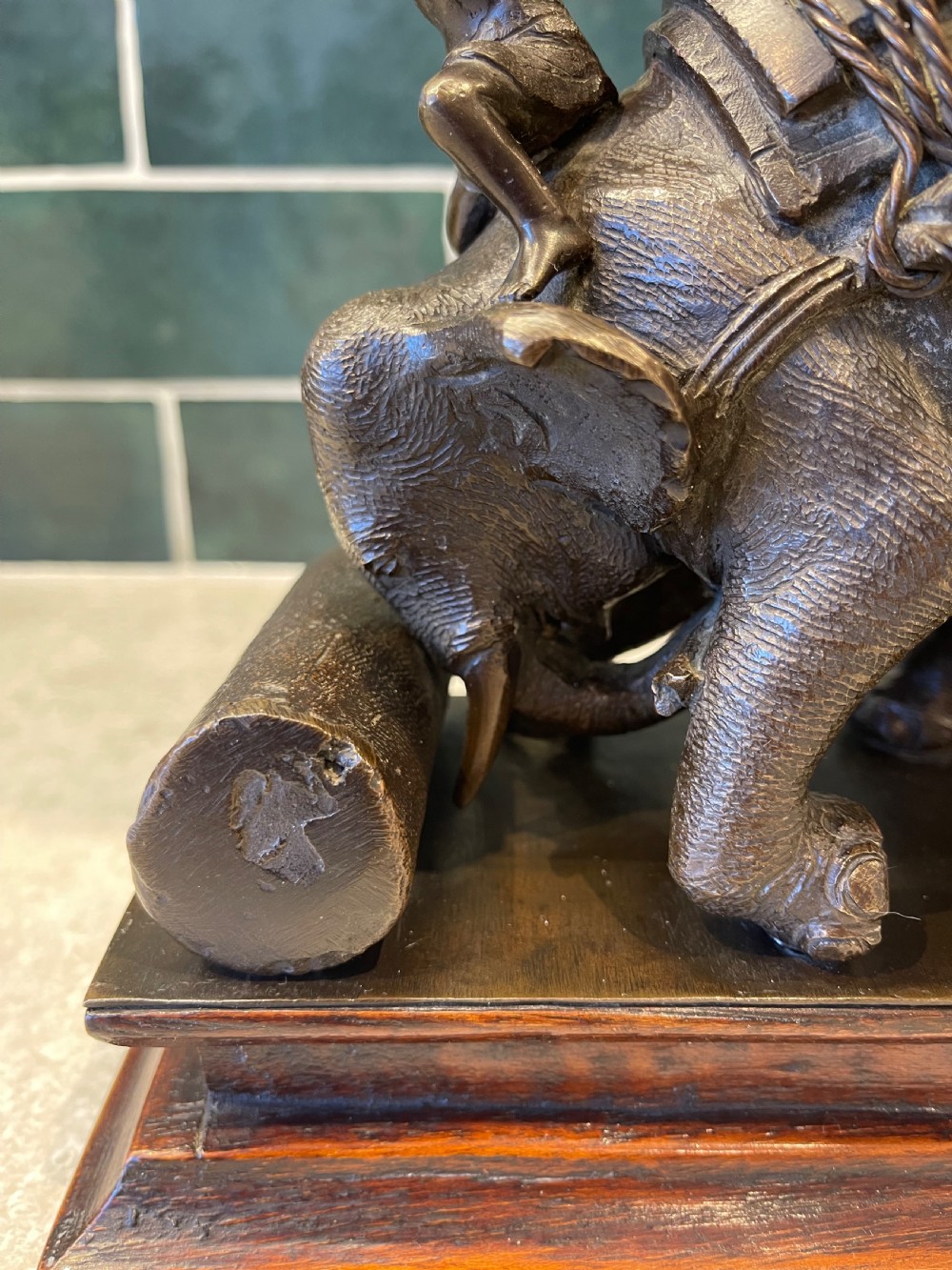 A patinated dark bronze sculpture of a working elephant pushing a log with a mahout riding its neck, mounted on a molded wooden base. View VIII