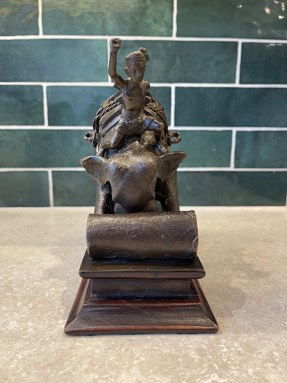 A patinated dark bronze sculpture of a working elephant pushing a log with a mahout riding its neck, mounted on a molded wooden base. View IX