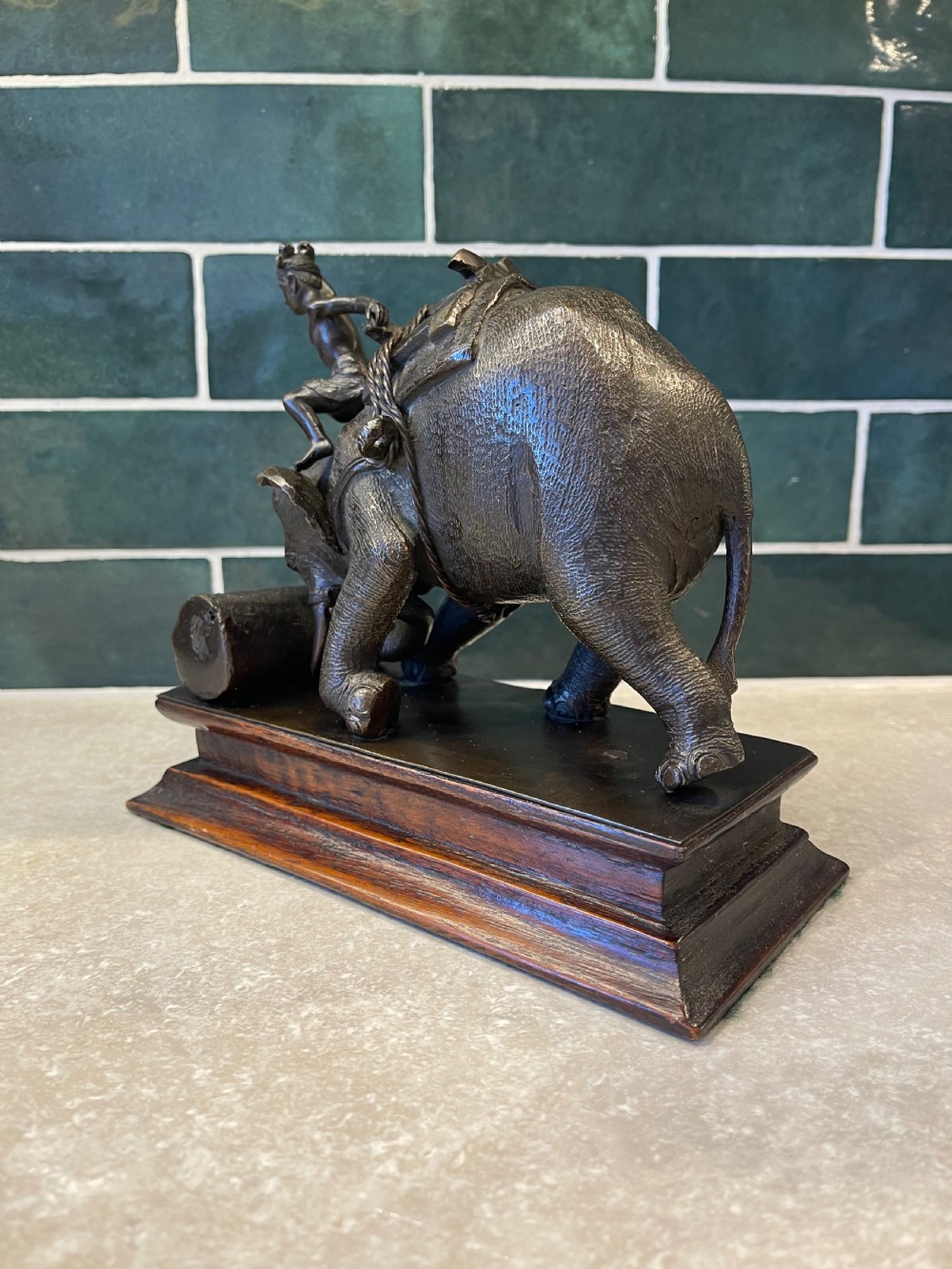 A patinated dark bronze sculpture of a working elephant pushing a log with a mahout riding its neck, mounted on a molded wooden base. View X