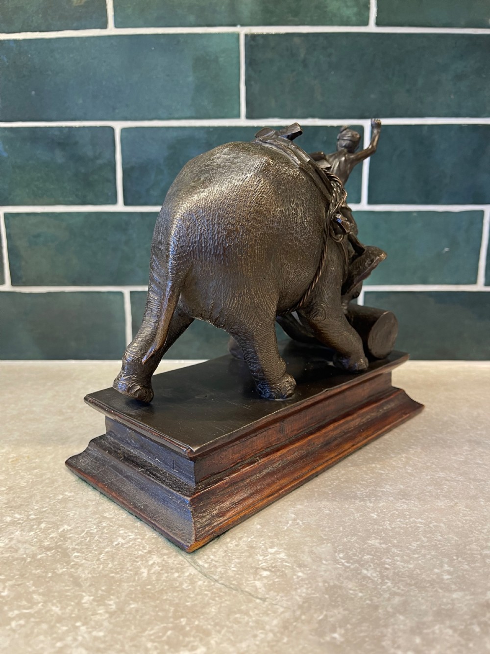 A patinated dark bronze sculpture of a working elephant pushing a log with a mahout riding its neck, mounted on a molded wooden base. View XI