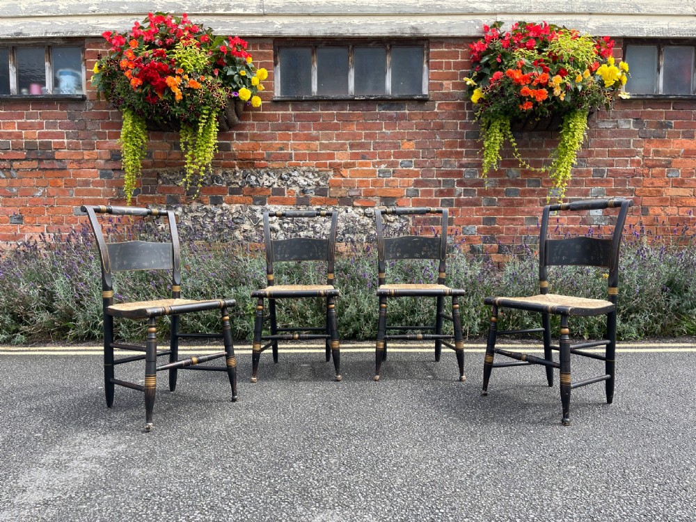 A set of four black-painted wooden side chairs featuring gold stencilled foliage, turned legs, horizontal back slats, and rush seats.