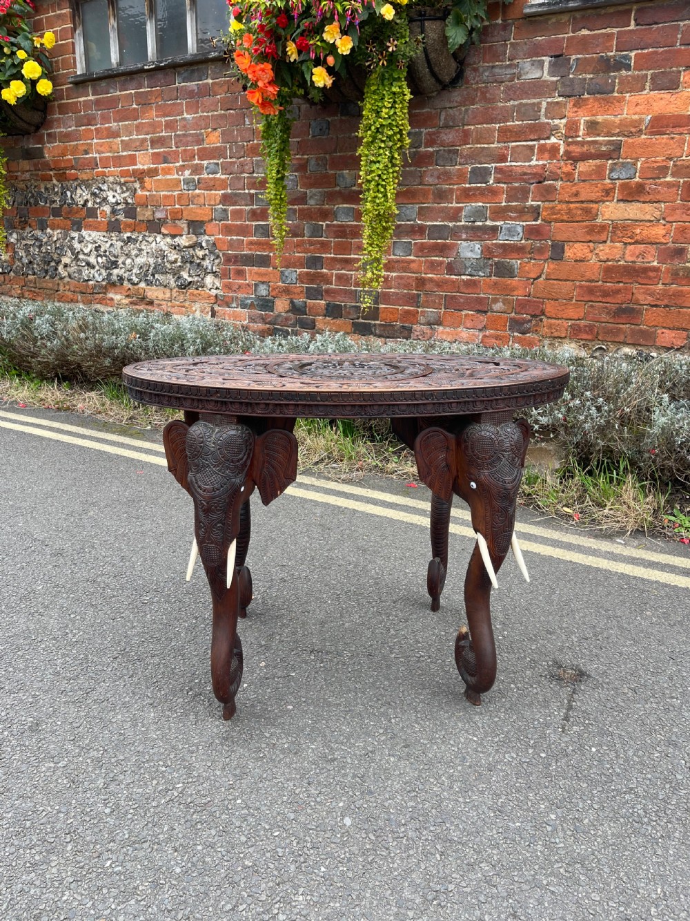 A circular dark carved wooden side table supported by three legs shaped like detailed elephant heads with trunks reaching to the floor.