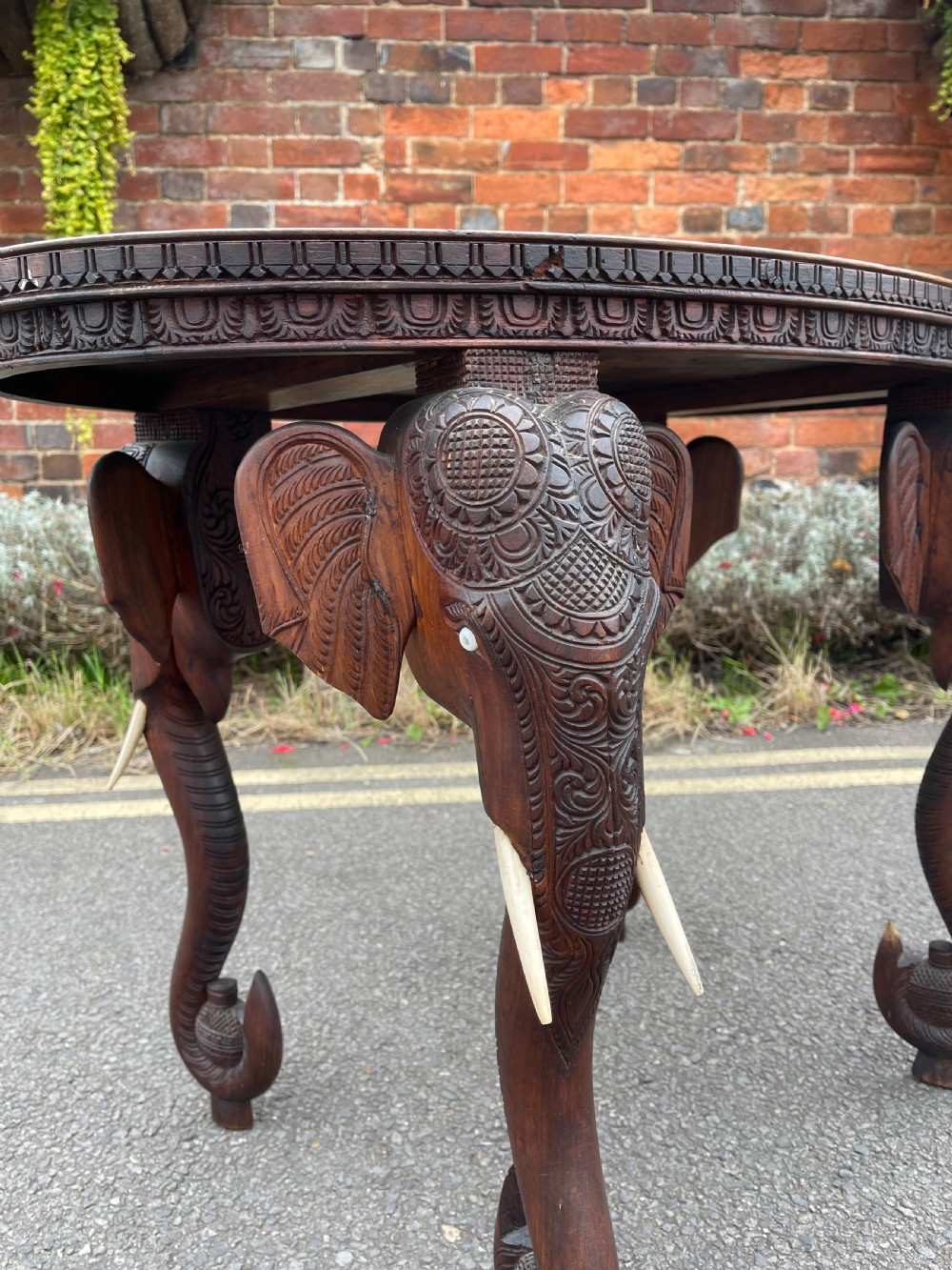 A circular dark carved wooden side table supported by three legs shaped like detailed elephant heads with trunks reaching to the floor. View II