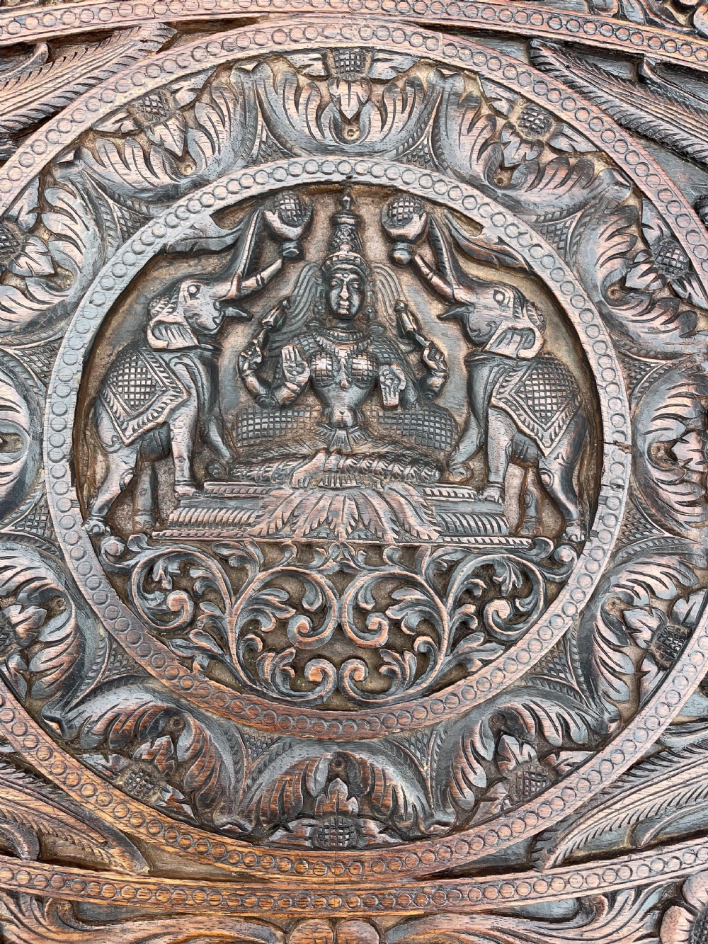 A circular dark carved wooden side table supported by three legs shaped like detailed elephant heads with trunks reaching to the floor. View III