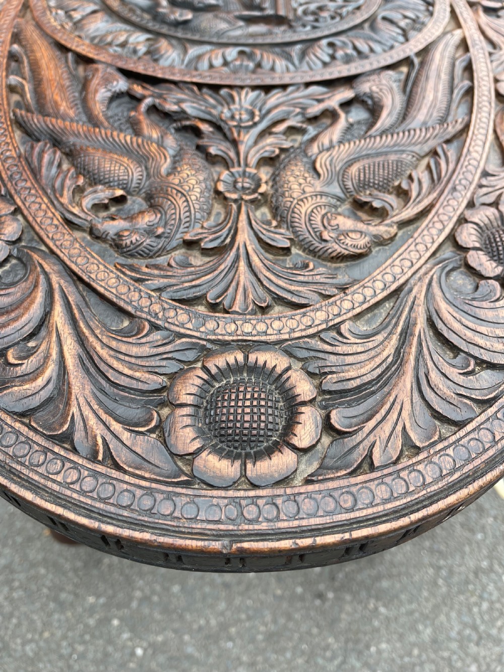 A circular dark carved wooden side table supported by three legs shaped like detailed elephant heads with trunks reaching to the floor. View V