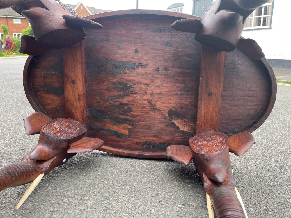 A circular dark carved wooden side table supported by three legs shaped like detailed elephant heads with trunks reaching to the floor. View VI