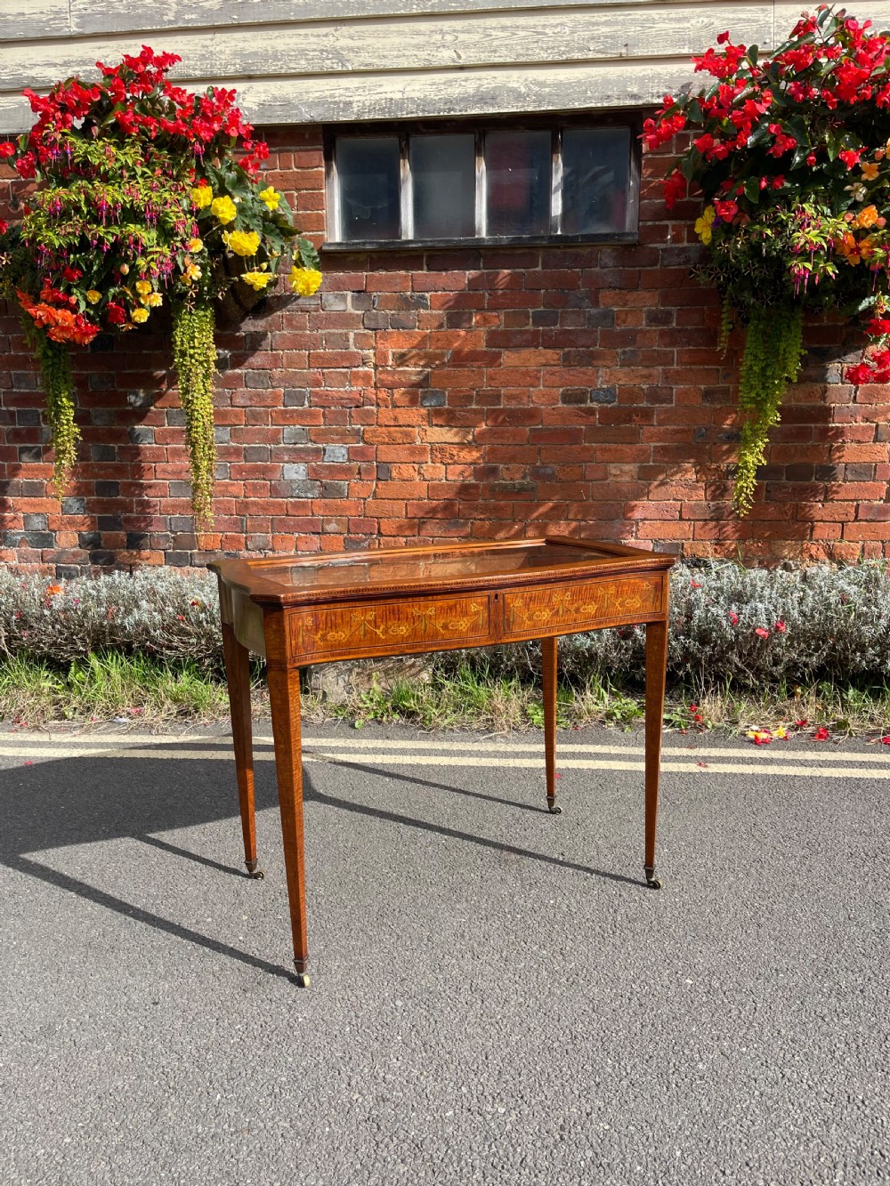 A late Victorian satinwood bijouterie table featuring a glazed display top, floral marquetry inlay along the frieze, and tapered legs with brass castors.