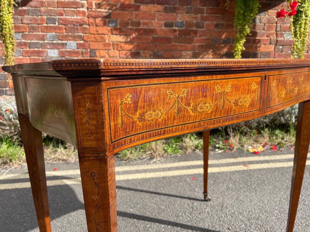 A late Victorian satinwood bijouterie table featuring a glazed display top, floral marquetry inlay along the frieze, and tapered legs with brass castors. View II