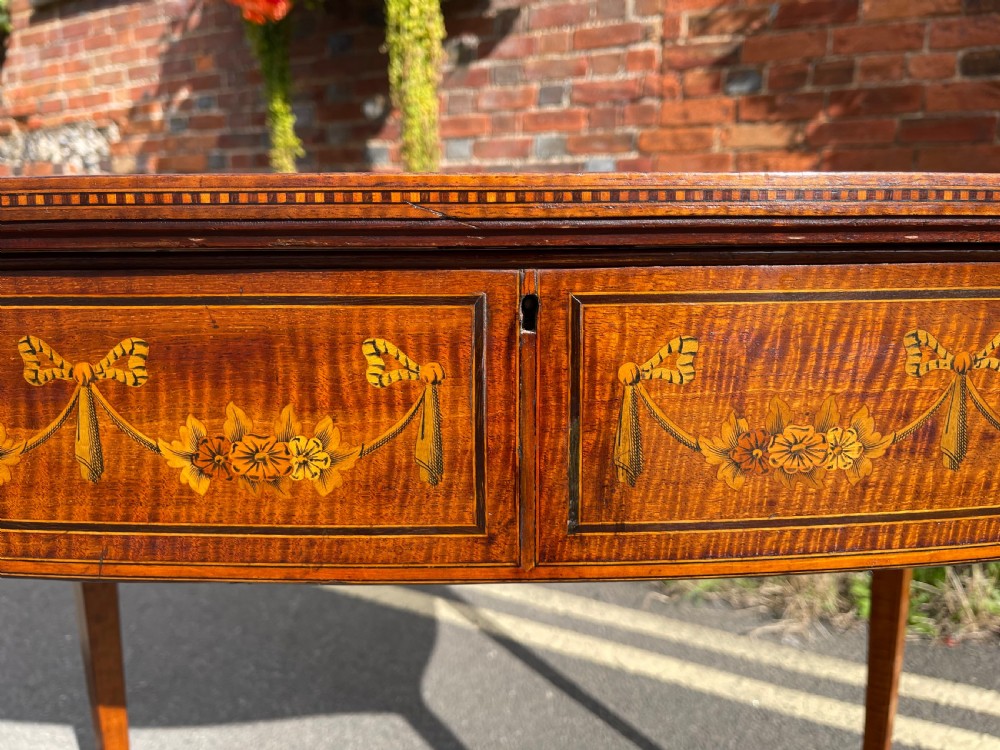 A late Victorian satinwood bijouterie table featuring a glazed display top, floral marquetry inlay along the frieze, and tapered legs with brass castors. View IV