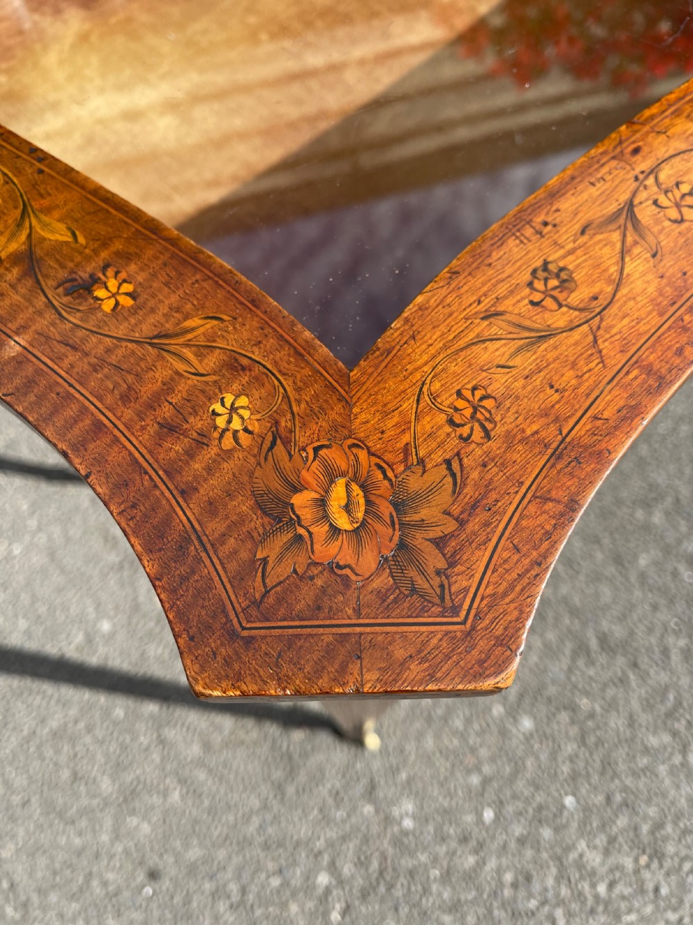 A late Victorian satinwood bijouterie table featuring a glazed display top, floral marquetry inlay along the frieze, and tapered legs with brass castors. View VI