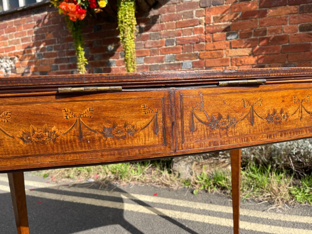 A late Victorian satinwood bijouterie table featuring a glazed display top, floral marquetry inlay along the frieze, and tapered legs with brass castors. View IX