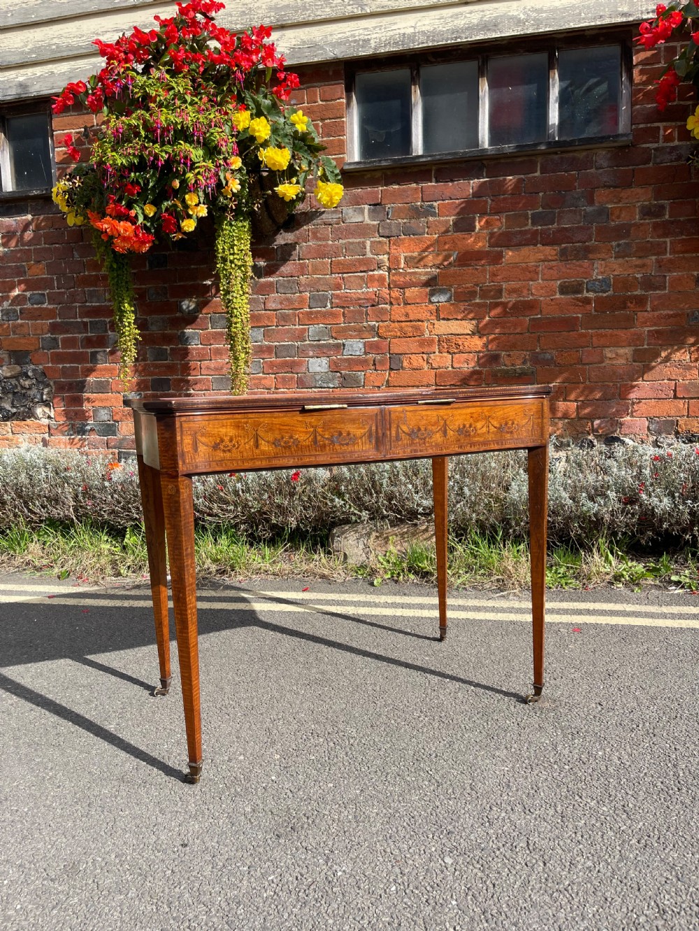 A late Victorian satinwood bijouterie table featuring a glazed display top, floral marquetry inlay along the frieze, and tapered legs with brass castors. View X