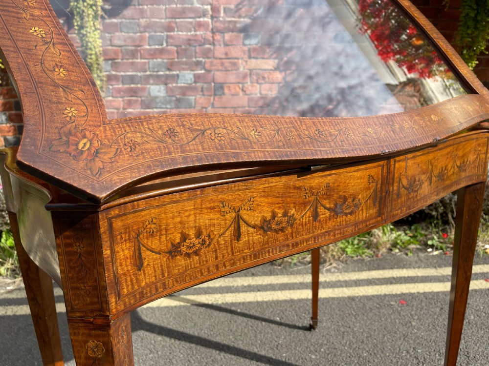 A late Victorian satinwood bijouterie table featuring a glazed display top, floral marquetry inlay along the frieze, and tapered legs with brass castors. View XIII