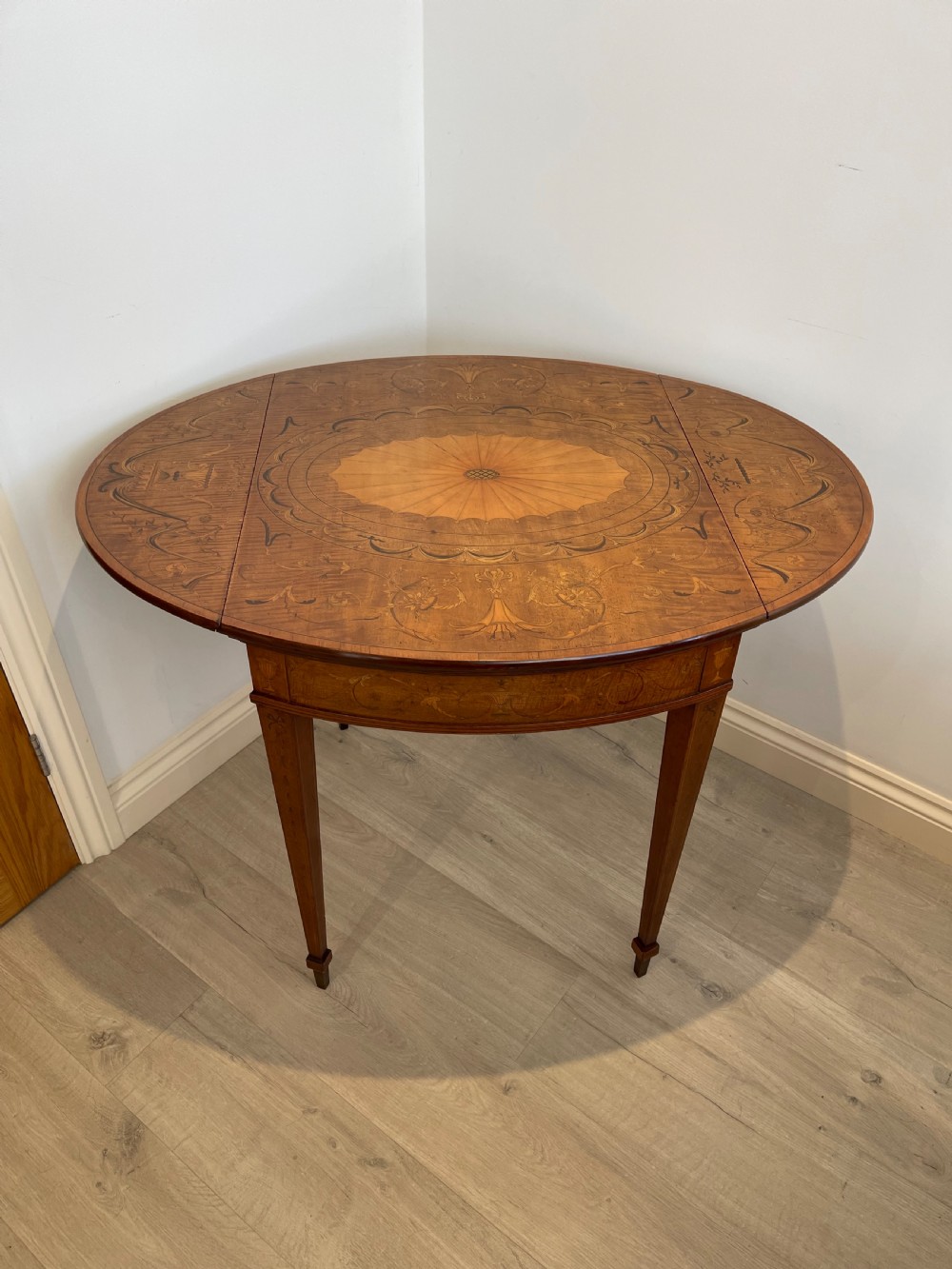 An oval antique mahogany Pembroke table with detailed marquetry inlay, drop leaves, and tapered legs.