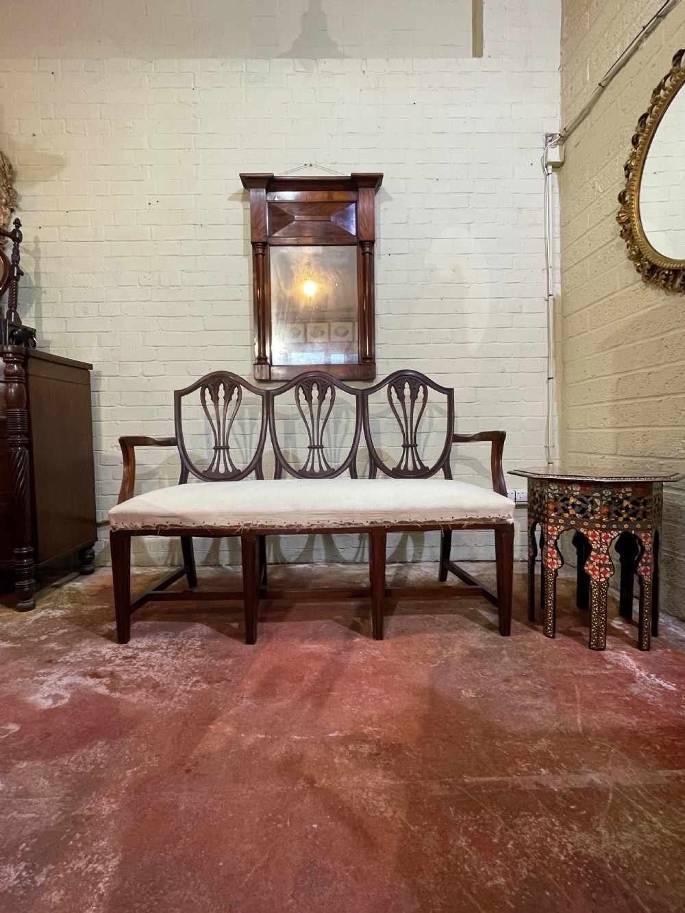 A dark mahogany triple chair-back settee with three pierced shield-shaped splats, swept armrests, square legs, and a neutral cream upholstered seat.