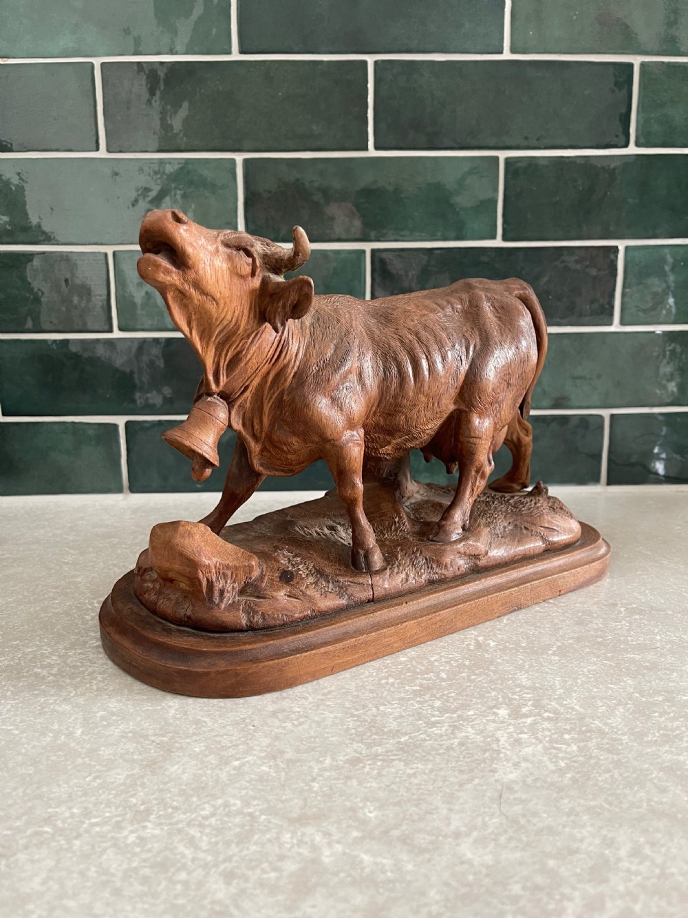 A detailed 19th-century carved wooden cow featuring an upturned head with a cowbell around its neck, standing on a carved naturalistic terrain base.