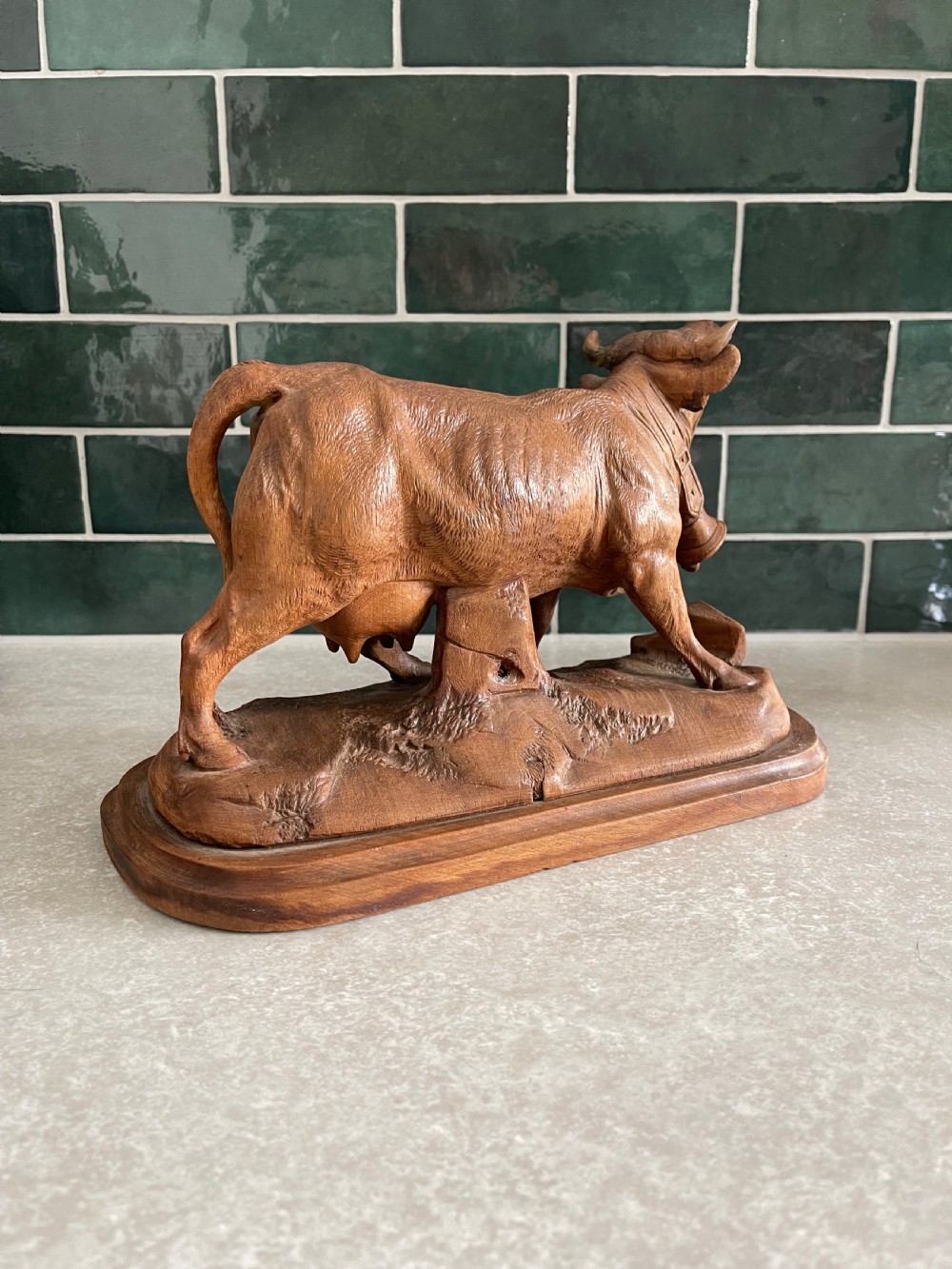 A detailed 19th-century carved wooden cow featuring an upturned head with a cowbell around its neck, standing on a carved naturalistic terrain base. View II